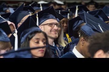 A graduate seated with his fellow graduates smiles up at the stage.